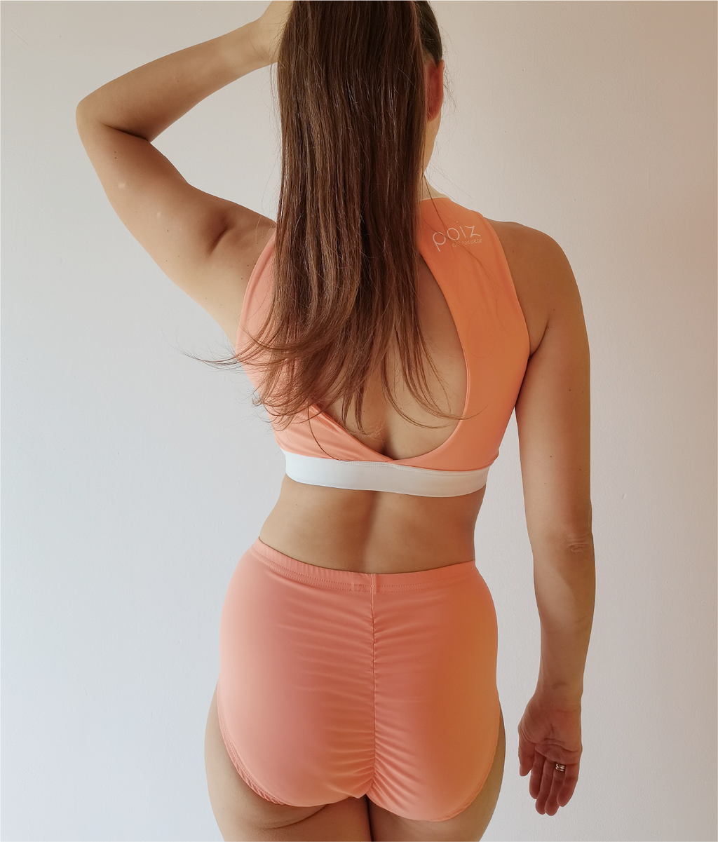Back shot Woman with brown hair and pony tail wearing orange-white fitness top with open back and shorts with scrunch butt